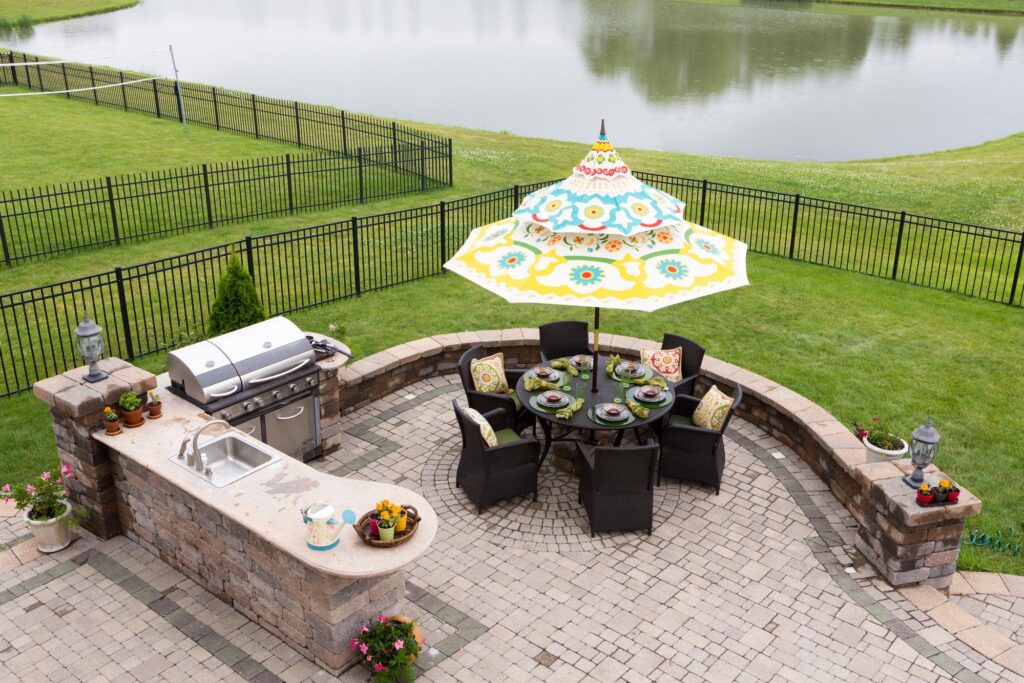 Outdoor patio dining set in an outdoor living space on a brick patio overlooking a tranquil lake and fenced green lawn with a table under a sunshade or umbrella laid ready for dinner, high angle view